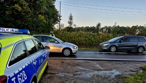 policjant stojący na drodze, kontroluje pojazd. Na pierwszym planie radiowóz