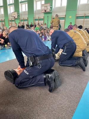 na sali policjanci i strażacy klęczą przed fantomami, przed nimi siedzą uczniowie
