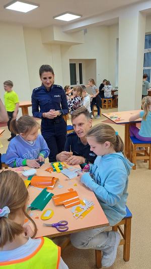 policjant klęczy przy stoliku przy którym siedzą dzieci. Na stole leżą odblaskowe fragmenty naklejek. Obok nich stoi policjantka