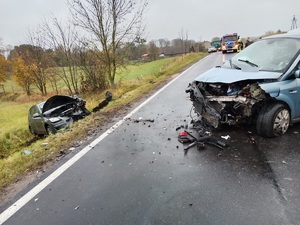 jeden samochód z rozbitym przodem stojący na drodze, kolejny rozbity samochód w rowie, w tle wóz straży pożarnej