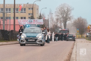radiowóz jadący drogą a za nim przemarsz ludzi z flagami ulicami Mikołajek