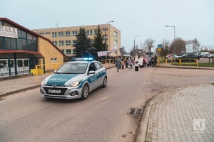 radiowóz jadący drogą a za nim przemarsz ludzi z flagami ulicami Mikołajek