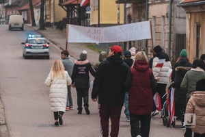 radiowóz jadący drogą a za nim przemarsz ludzi z flagami ulicami Mikołajek