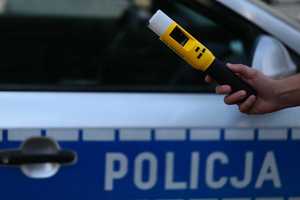 Obraz przedstawia rękę policjanta trzymająca urządzenie do badania stanu trzeźwości kierujących. W tle widoczny jest policyjny radiowóz.