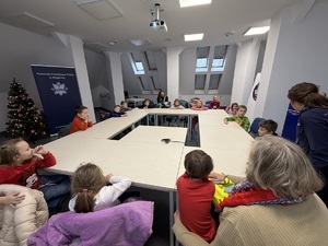 dzieci siedzą przy stole na sali odpraw, przy nich stoi policjantka