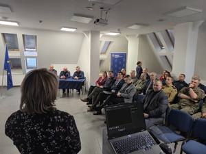 osoby siedzące na sali odpraw a przed nimi stoi kobieta