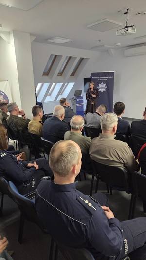 osoby siedzące na sali odpraw a przed nimi stoi kobieta