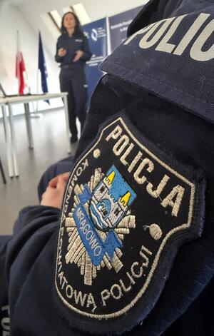 Zdjęcie przedstawia naszywkę z logiem Komendy Powiatowej Policji w Mrągowie, a w tle widoczna jest policjantka prowadząca wykład.