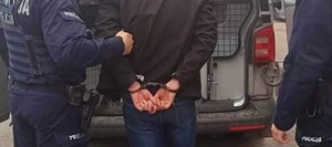 Zdjęcie przedstawia mężczyznę z kajdankami założonymi na ręce trzymane z tyłu, który jest osadzany przez policjantów w radiowozie.