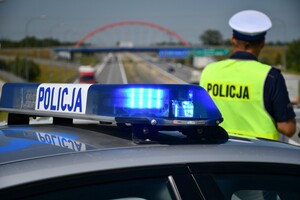 Zdjęcie przedstawia policjanta i radiowóz na tle drogi.