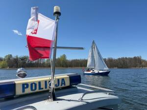 Zdjęcie przedstawia łódź policyjną, a w tle widać żaglówkę oraz jezioro Czos.