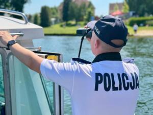 Zdjęcie przedstawia policjanta, znajdującego się na łodzi policyjnej. Policjant patrzy przez lornetkę.