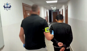 Zdjęcie przedstawia nieumundurowanego policjanta prowadzącego mężczyznę w kajdankach.
