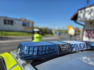 Zdjęcie przedstawia sygnały radiowozu z napisem POLICJA oraz stojącego w oddali policjanta ruchu drogowego w kamizelce odblaskowej.