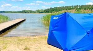 Zdjęcie przedstawia rozstawiony na plaży parawan z napisem POLICJA. W tle widać pomost oraz jezioro.