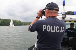 Zdjęcie przedstawia policjanta stojącego na łodzi policyjnej, który obserwuje przez lornetkę sytuację na jeziorze.
